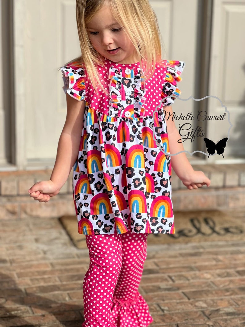 Puede incluir: Una ni&ntilde;a peque&ntilde;a lleva un vestido de lunares rosa y blanco con un dise&ntilde;o de arco&iacute;ris y estampado de leopardo. El vestido tiene un borde de volantes y un cierre de bot&oacute;n. Tambi&eacute;n lleva pantalones de lunares rosa con un borde de volantes en la parte inferior.