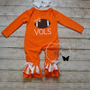 May include: Orange baby onesie with a white football design and the word "VOLS" on the front. The onesie has white ruffle trim on the legs and a white bow on the football.