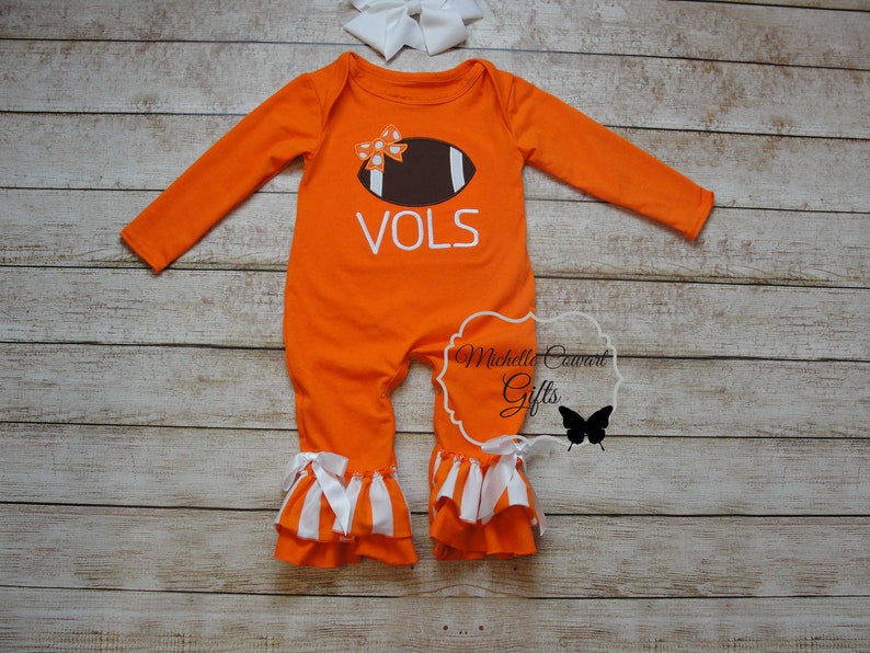 May include: Orange baby onesie with a white football design and the word "VOLS" on the front. The onesie has white ruffle trim on the legs and a white bow on the football.