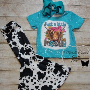 Pode incluir: Um conjunto de camisa turquesa de manga curta e cal&ccedil;as com estampa de vaca. A camisa apresenta um gr&aacute;fico de uma vaca Highland com o texto "Just a little moody". Inclui um grande la&ccedil;o turquesa com centro de strass.
