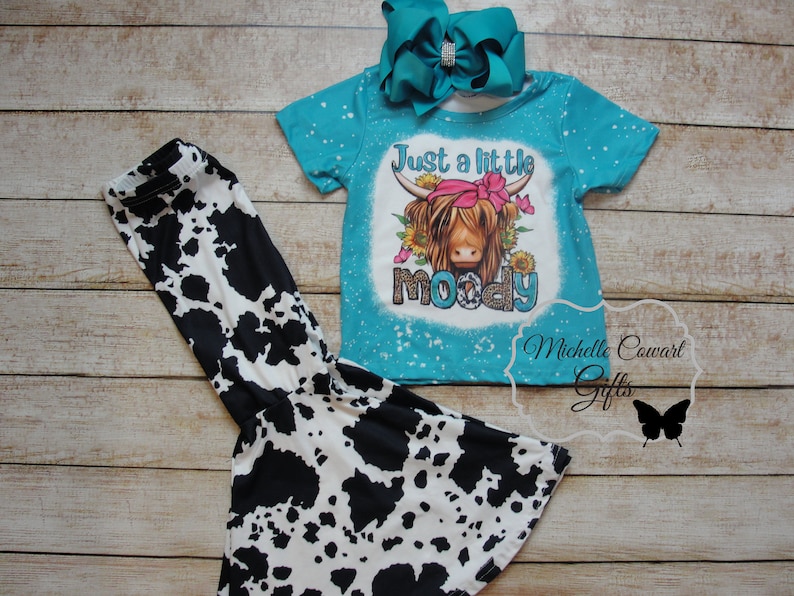 Pode incluir: Um conjunto de camisa turquesa de manga curta e cal&ccedil;as com estampa de vaca. A camisa apresenta um gr&aacute;fico de uma vaca Highland com o texto "Just a little moody". Inclui um grande la&ccedil;o turquesa com centro de strass.