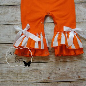 May include: Orange and white striped baby romper with white satin bows and ruffles. The romper has snap closures on the crotch.