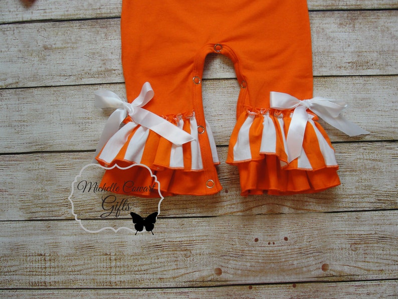 May include: Orange and white striped baby romper with white satin bows and ruffles. The romper has snap closures on the crotch.