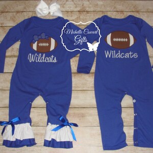 May include: Two blue long-sleeved baby onesies with white ruffles at the bottom. One onesie has a white embroidered football with the text "Wildcats" and the other onesie has a white embroidered football with the text "Wildcats".