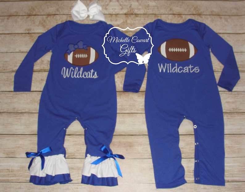 May include: Two blue long-sleeved baby onesies with white ruffles at the bottom. One onesie has a white embroidered football with the text "Wildcats" and the other onesie has a white embroidered football with the text "Wildcats".