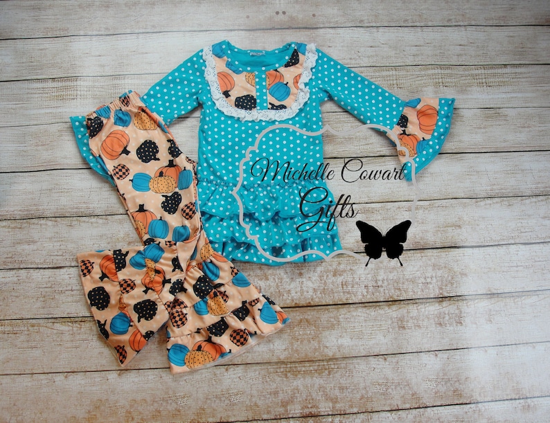 Puede incluir: Conjunto de top y pantal&oacute;n turquesa con lunares. El top tiene mangas largas con pu&ntilde;os acampanados y un dobladillo con volantes. El pantal&oacute;n presenta un estampado de calabaza. El conjunto incluye el texto "Michelle Cowart Gifts" y una silueta de mariposa.