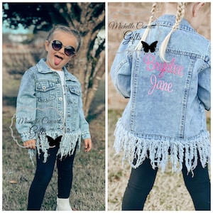 May include: A light blue denim jacket with frayed edges and a pink embroidered name "Brynnlee Jane" on the back. The jacket has a butterfly design on the front.