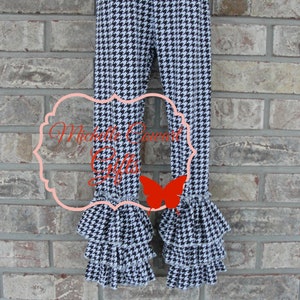 May include: Black and white houndstooth patterned pants with ruffled hems. The pants have a fitted design and are displayed against a brick wall. The text "Michelle Cowart Gifts" is visible on the pants.