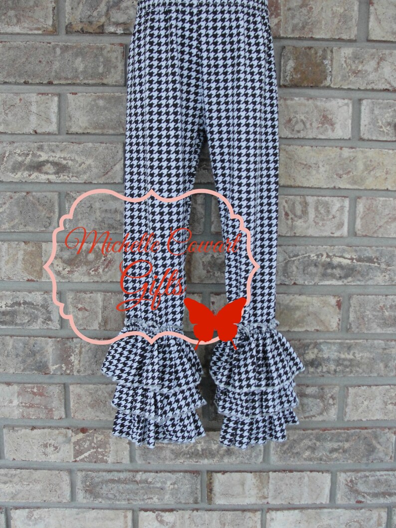 May include: Black and white houndstooth patterned pants with ruffled hems. The pants have a fitted design and are displayed against a brick wall. The text "Michelle Cowart Gifts" is visible on the pants.