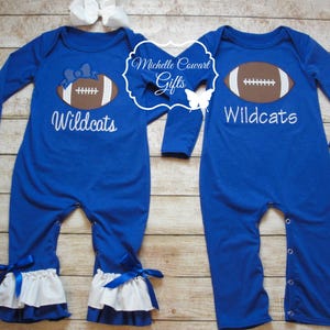 May include: Two blue long-sleeved baby onesies with white ruffles at the bottom. One onesie has a brown football with a white bow on top and the text "Wildcats". The other onesie has a brown football and the text "Wildcats".