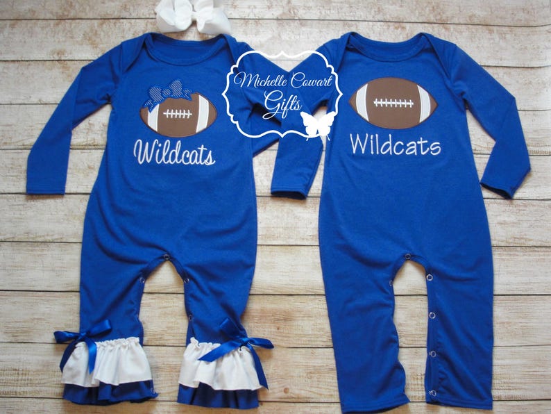 May include: Two blue long-sleeved baby onesies with white ruffles at the bottom. One onesie has a brown football with a white bow on top and the text "Wildcats". The other onesie has a brown football and the text "Wildcats".