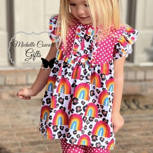 Puede incluir: Una blusa de lunares rosa y blanca con una falda de estampado de arco&iacute;ris y leopardo. La blusa tiene cierre de bot&oacute;n y mangas con volantes. La falda tiene un dobladillo con volantes. El atuendo lo lleva una ni&ntilde;a peque&ntilde;a.