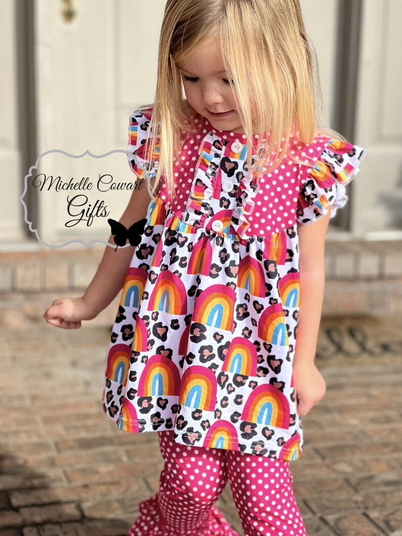 Puede incluir: Una blusa de lunares rosa y blanca con una falda de estampado de arco&iacute;ris y leopardo. La blusa tiene cierre de bot&oacute;n y mangas con volantes. La falda tiene un dobladillo con volantes. El atuendo lo lleva una ni&ntilde;a peque&ntilde;a.
