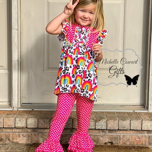 Puede incluir: Una ni&ntilde;a peque&ntilde;a con un pantal&oacute;n de volantes de lunares rosa y blanco con una blusa de estampado de arco&iacute;ris y leopardo. La blusa tiene un cuello de volantes y mangas cortas. La ni&ntilde;a est&aacute; de pie frente a una puerta con una alfombra de bienvenida que dice "hello".