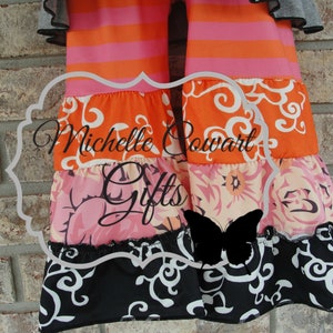 May include: A colorful tiered garment with orange and pink stripes, floral patterns, and a black and white scroll design. The text "Michelle Powart Gifts" is visible. The garment has ruffled edges and a butterfly silhouette.