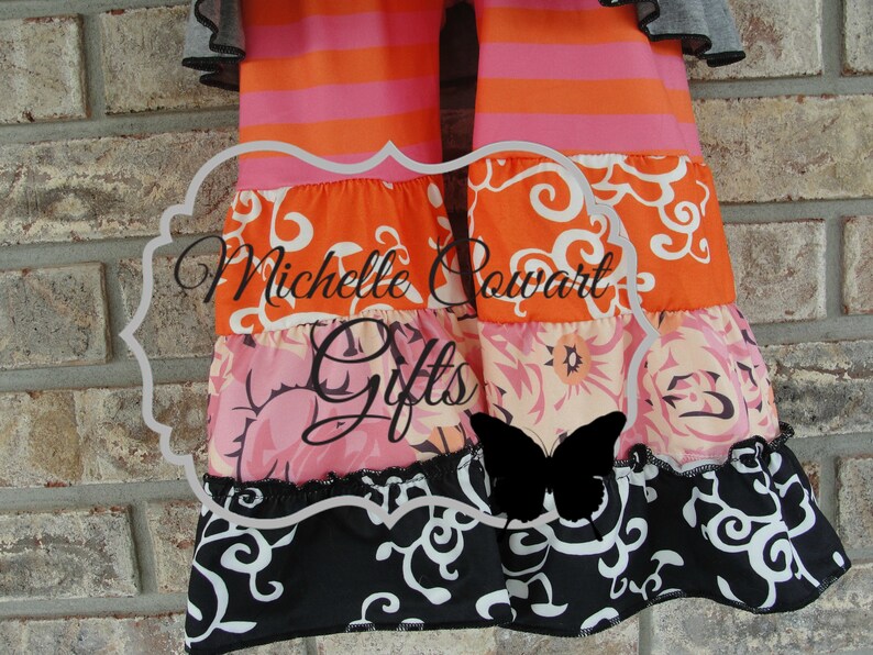 May include: A colorful tiered garment with orange and pink stripes, floral patterns, and a black and white scroll design. The text "Michelle Powart Gifts" is visible. The garment has ruffled edges and a butterfly silhouette.