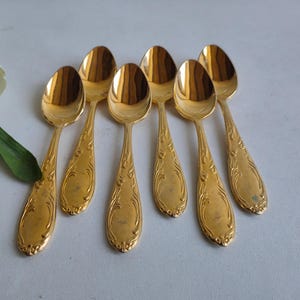 May include: A set of seven gold-colored spoons with ornate, engraved handles. The spoons are arranged on a white surface, with a sprig of white flowers and green leaves in the upper left corner. The spoons are likely for serving or dessert.