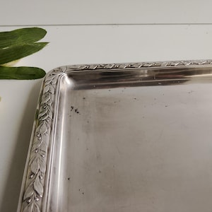 May include: A rectangular silver-colored tray with a decorative leaf and berry border. The tray has a reflective surface and rounded corners. The tray is likely made of metal and is suitable for serving or display.