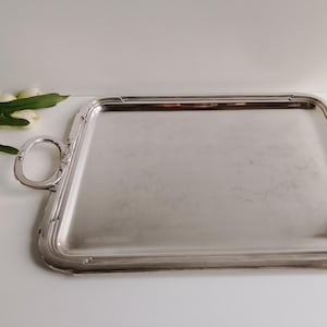 Op de afbeelding: Een rechthoekig zilveren serveerblad met afgeronde handgrepen. De lade heeft een reflecterend oppervlak en een decoratieve rand. De handgrepen zijn ovaal van vorm en aan de zijkanten van de lade bevestigd. De lade wordt waarschijnlijk gebruikt voor het serveren van eten of drinken.