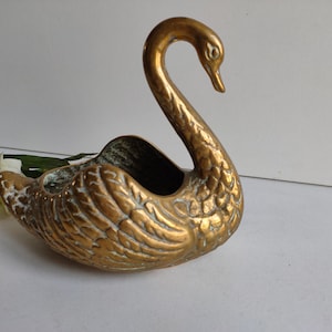 May include: A gold swan-shaped planter with a detailed design. The planter is made of brass and has a worn, antique look.