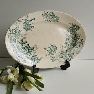 Puede incluir: Plato ovalado antiguo con fondo crema y diseño floral turquesa. El plato está sostenido por un soporte negro, con un ramo de flores blancas en primer plano.