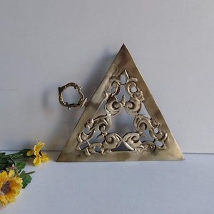 May include: A brass triangle with a decorative cutout design. The triangle has a small loop handle on one side.