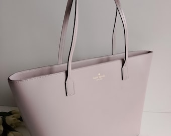 May include: A light purple tote bag with two long handles. The bag has a rectangular shape and a small logo on the front. The handles are thin and made of the same material as the bag. The bag is made of a smooth material.