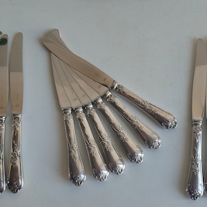 May include: A selection of silver knives with decorative, floral-patterned handles. The blades are polished and reflective. The knives are displayed on a light surface, possibly for a formal dinner. The set includes several knives.