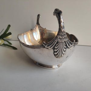 May include: A silver-toned decorative basket with a handle featuring an ornate bird design. The basket has a rounded shape with a detailed rim and a pedestal base. The handle is intricately designed, adding to the vintage aesthetic.