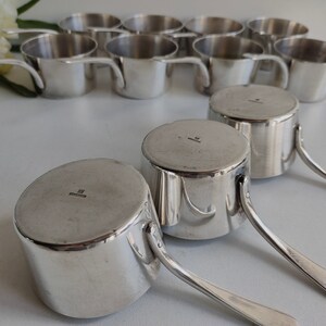 May include: A collection of small, shiny silver metal saucepans with handles. The saucepans are cylindrical with a flat bottom and a small handle attached to the side. The image shows several saucepans, some in the background and some in the foreground.