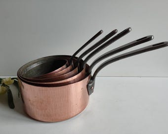 May include: Set of five copper saucepans with black handles. The saucepans are nested together, with the smallest saucepan on top. The saucepans are all the same shape and size, and they have a hammered finish.