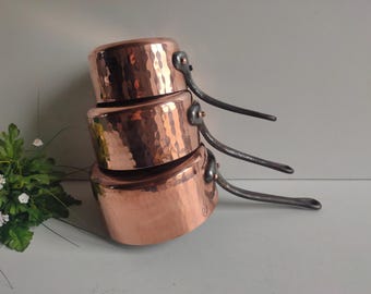 May include: A set of three copper pots with hammered surfaces and black iron handles. The pots are stacked on top of each other, with the smallest pot on top.