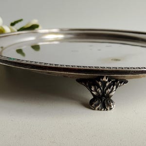 Pode incluir: Uma bandeja redonda prateada com uma borda decorativa e pés ornamentados em estilo floral. A bandeja é reflexiva, e os detalhes da borda são visíveis. Algumas flores brancas estão no fundo.