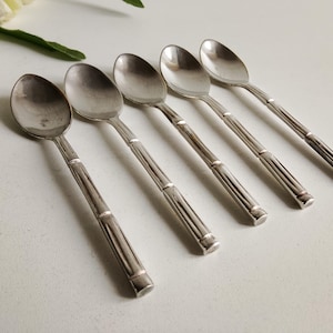 May include: Five silver spoons with a bamboo-style handle design are arranged on a white surface. The spoons have a classic oval bowl shape and a polished finish. The spoons are likely for serving or eating.