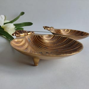 May include: Two antique gold-colored seashell-shaped dishes. Each dish has a textured, ridged surface and is supported by three small feet. The dishes have a decorative floral accent at the top.
