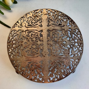 Puede incluir: Un salvamanteles de metal plateado con un diseño floral decorativo e intrincado. El salvamanteles es redondo y tiene un patrón en forma de cruz en el centro.