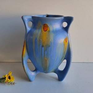 Peut inclure: Un vase en céramique bleu avec des accents jaunes et orange. Le vase a une forme unique avec trois pieds et deux poignées.