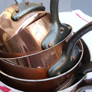May include: A collection of copper saucepans and frying pans with dark metal handles. The cookware has a polished, reflective surface. The handles are attached with rivets. The pans are likely for cooking and food preparation.