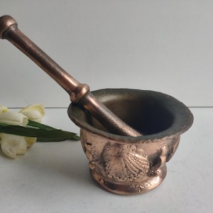 May include: A copper mortar and pestle with an ornate design. The mortar is round and has a textured surface. The pestle is long and has a rounded end.