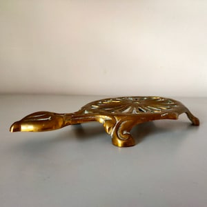 Pode incluir: Um descanso de panela de metal dourado em forma de tartaruga com um design decorativo vazado. O descanso de panela é projetado para segurar pratos quentes.