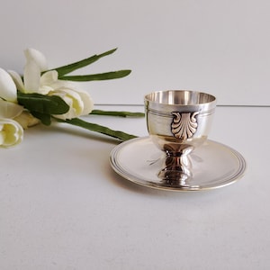 May include: A silver-toned metal egg cup with a matching saucer. The cup features a decorative shell-like motif on its side. The saucer has a smooth, reflective surface with a thin circular border detail. The set is polished and metallic.