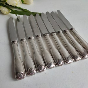 May include: A collection of ten silver-coloured knives. Each knife features a decorative shell design on the handle and a smooth, shiny blade. The knives are arranged in a row, suitable for a formal dining occasion.