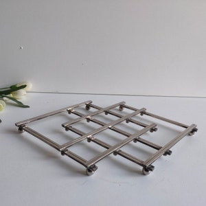 Peut inclure: Un dessous de plat en métal argenté avec un design géométrique. Le dessous de plat est composé de cadres rectangulaires interconnectés, soutenus par de petits pieds sphériques. Il est conçu pour protéger les surfaces des ustensiles de cuisine chauds.