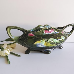 May include: A dark green ceramic planter with a glossy finish, featuring a floral design in pink, yellow, and blue. The planter has two handles and rests on three small feet. Decorative and vintage.