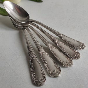 Può includere: Set di sei cucchiaini d'argento con manici decorati e dettagliati. I cucchiaini sono disposti a ventaglio, con le punte dei cucchiaini rivolte verso lo spettatore.