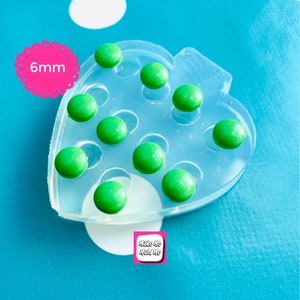 May include: A clear silicone mold shaped like a heart with 12 round cavities. The mold is filled with 6 mm green resin cabochons. The mold is labeled "Make Me Mold Me".