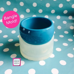 Peut inclure: Un moule à bracelet en silicone transparent avec un bracelet bleu à l'intérieur. Le moule est sur un fond bleu et blanc à pois. Le texte "Bangle Mold!" est en rose sur fond blanc. Le texte "Make Me Mold Me" est en blanc sur fond rose.