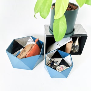Peut inclure: Deux contenants de rangement géométriques bleus avec différents objets à l'intérieur. Le plus grand contient une pochette en cuir marron et un stylo bleu. Le plus petit contient un jeu de clés et un petit récipient rond à motif floral.