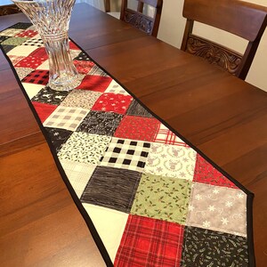 May include: A patchwork table runner with a black border, featuring squares of red, green, black, and white fabrics. Patterns include plaid, snowflakes, and holly. A clear glass vase sits on the runner, which is placed on a wooden table.
