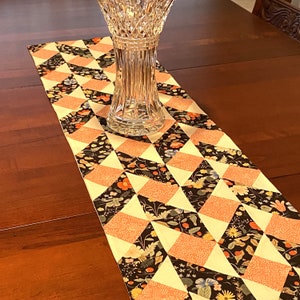 Op de afbeelding: Een decoratieve tafelloper met een geometrisch patroon van perzik, zwarte en crèmekleurige ruitvormen. De ruiten zijn gevuld met bloemen- en stippenontwerpen. Een heldere kristallen vaas staat op de loper, die op een donkere houten tafel ligt.